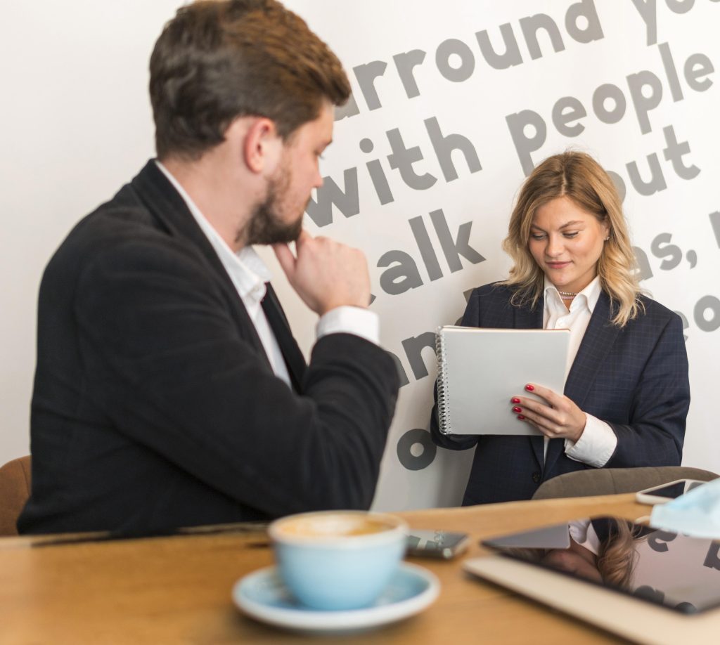 Two people in an office setting, sitting at a table with a notebook, coffee, and tablet. The woman appears to be referencing notes while the man listens thoughtfully.