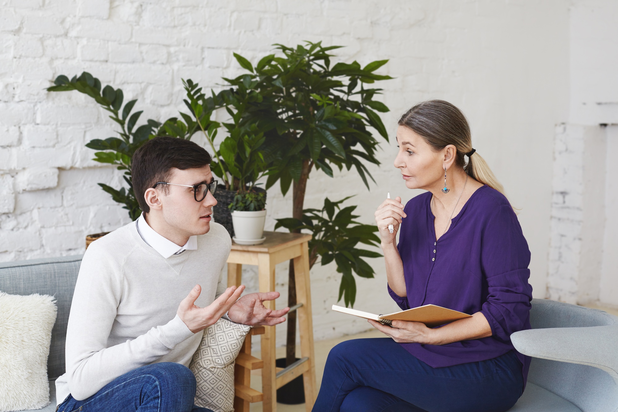 man and a woman are sitting on a couch, both looking at an open book. They appear engaged and are smiling, surrounded by indoor plants and furniture. The setting is cozy and homely.