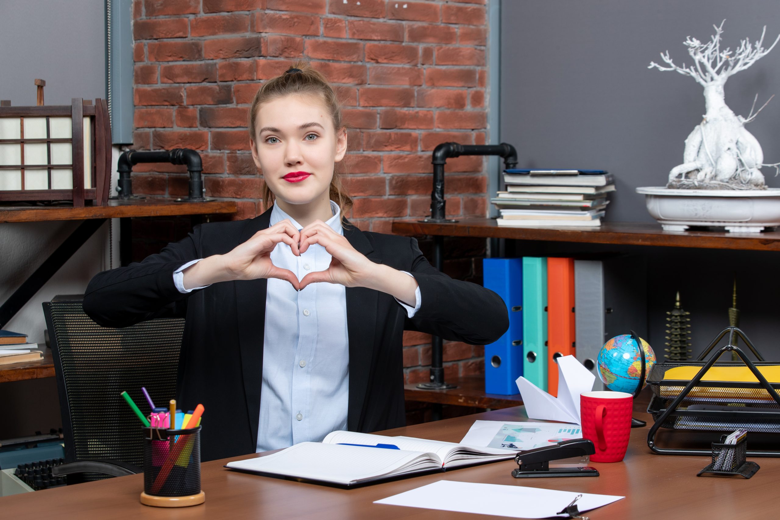 A lady in a black blazer forms a heart shape with hands while sitting at a desk cluttered with office supplies, a globe, and documents