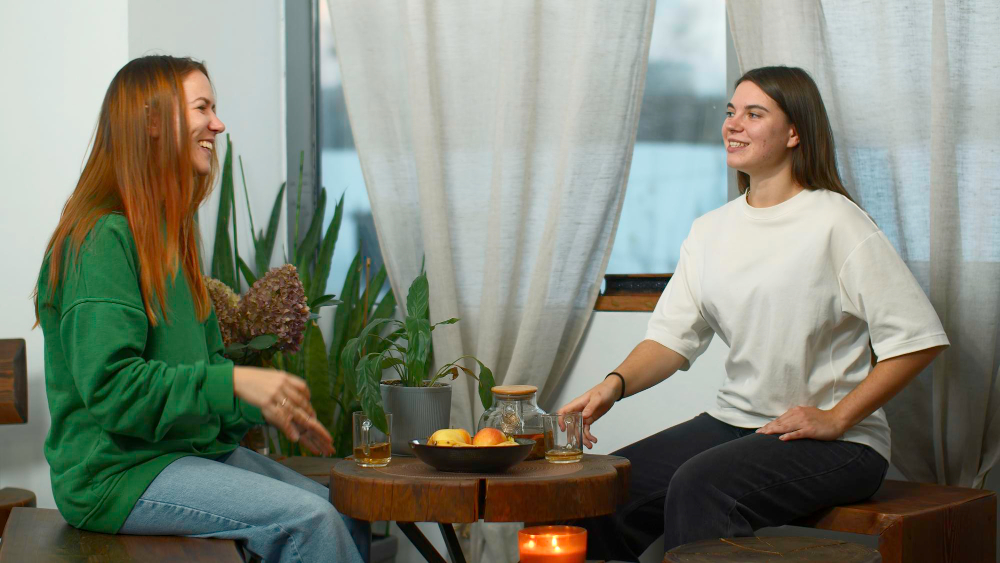 Two women enjoying a friendly conversation in a cozy living room, sitting at a wooden table with plants, oranges, and a candle. The warm atmosphere suggests relaxation and connection, ideal for lifestyle and social interaction themes.