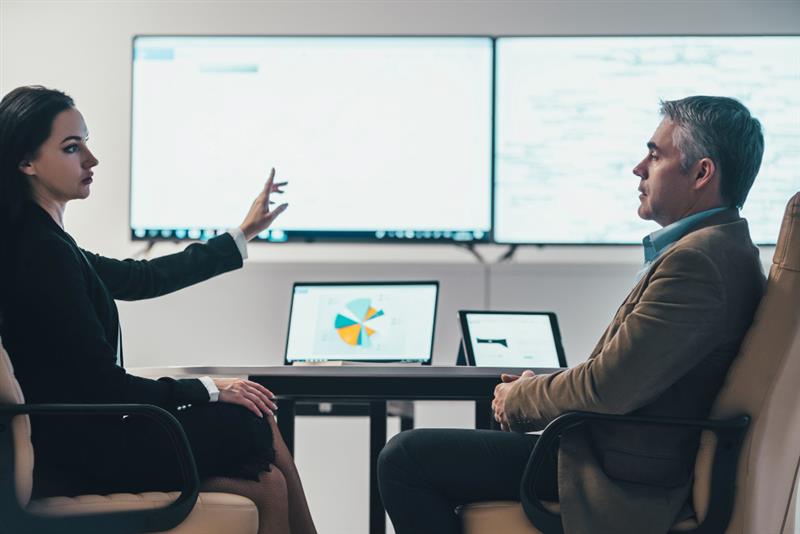 Two professionals in a meeting with laptops and presentation screens in the background.