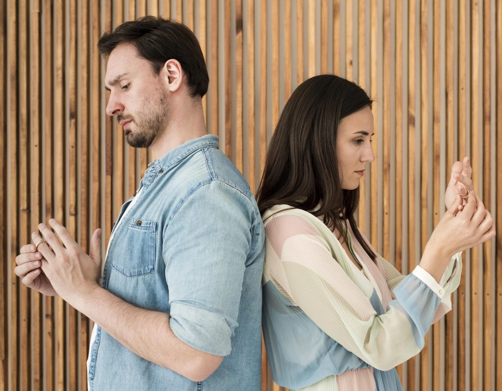 A couple standing back to back in front of a wooden wall, both looking at their wedding rings, symbolizing relationship issues or divorce. The man is wearing a denim shirt, and the woman is dressed in a colorful blouse.